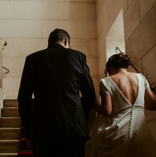 Central PA bride with an open back, short-sleeved wedding dress with lace detail and button going down the skirt walking with their backs to the camera up stone stairs with a groom in a dark suit.