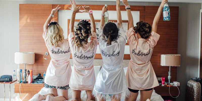 Four women wearing matching satin robes — three in blush pink that say “Bridesmaid” and one in white that says “Bride” — kneel on a hotel bed with their arms raised, celebrating together before the bachelorette festivities.