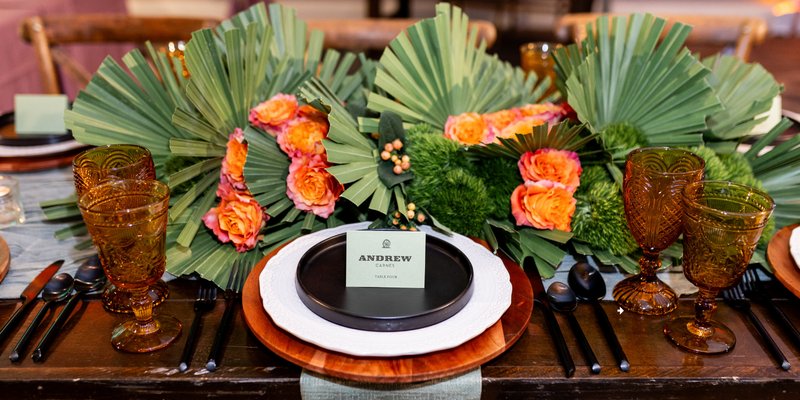 A rustic western-inspired wedding tablescape featuring amber glassware, black flatware, and layered plates with a green place card reading “Andrew.” The centerpiece includes bold orange roses, textured greenery, and fan-shaped palm leaves, creating a vibrant Oklahoma!-inspired look.