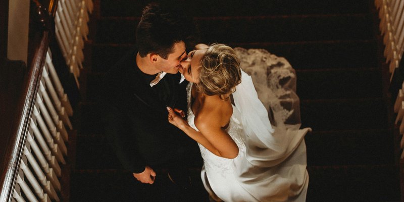 Overhead view of a bride and groom kissing on a staircase, with the bride in a white lace gown and veil and the groom in a black suit during their wedding.