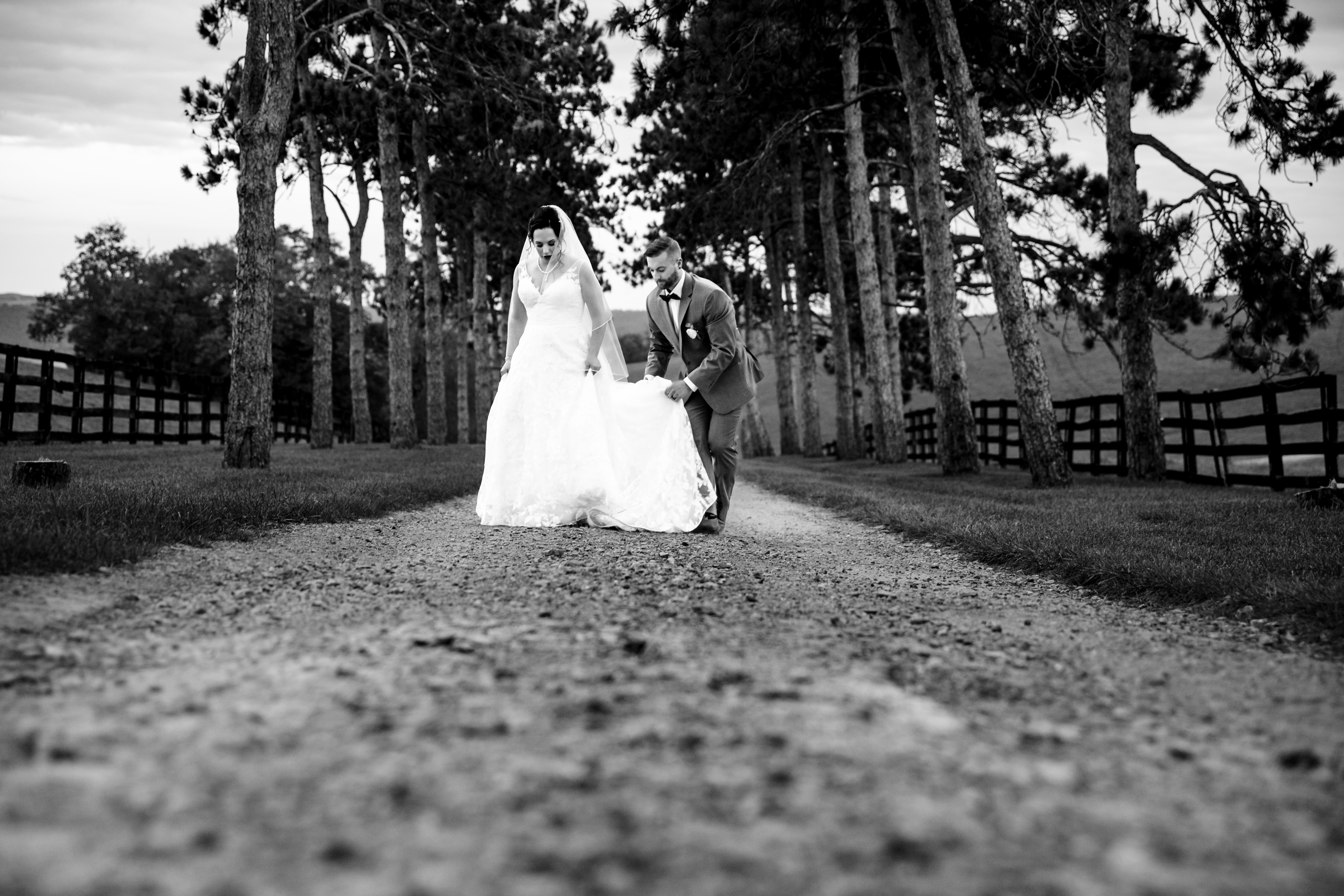 Romantic Barn Wedding Portrait Groom helps adjust the bride's dress as they walk down a tree-lined gravel lane at a Pennsylvania wedding venue.