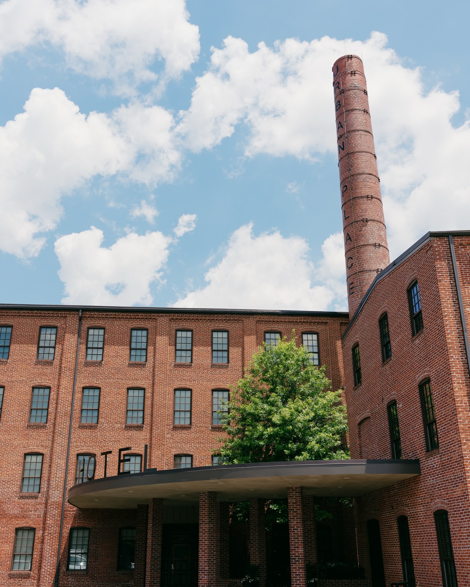 Cork Factory Hotel Exterior in Lancaster, Pennsylvania Historic red-brick boutique hotel with tall smokestack and black window frames under a bright blue sky with clouds.