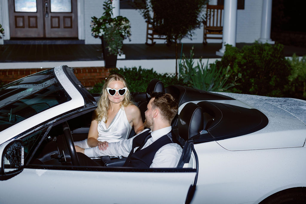 Bride wearing white heart sunglasses sitting in the front seat of a white convertible next to the groom