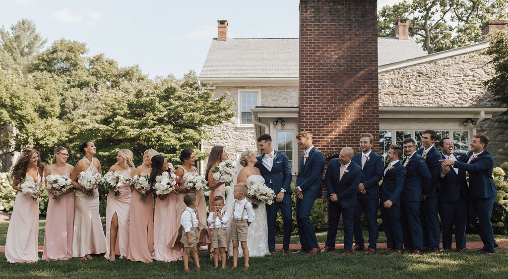 Bridesmaids in pink long dresses holding bouquets of white flowers and groomsmen in navy suits standing on either side of a bride and groom