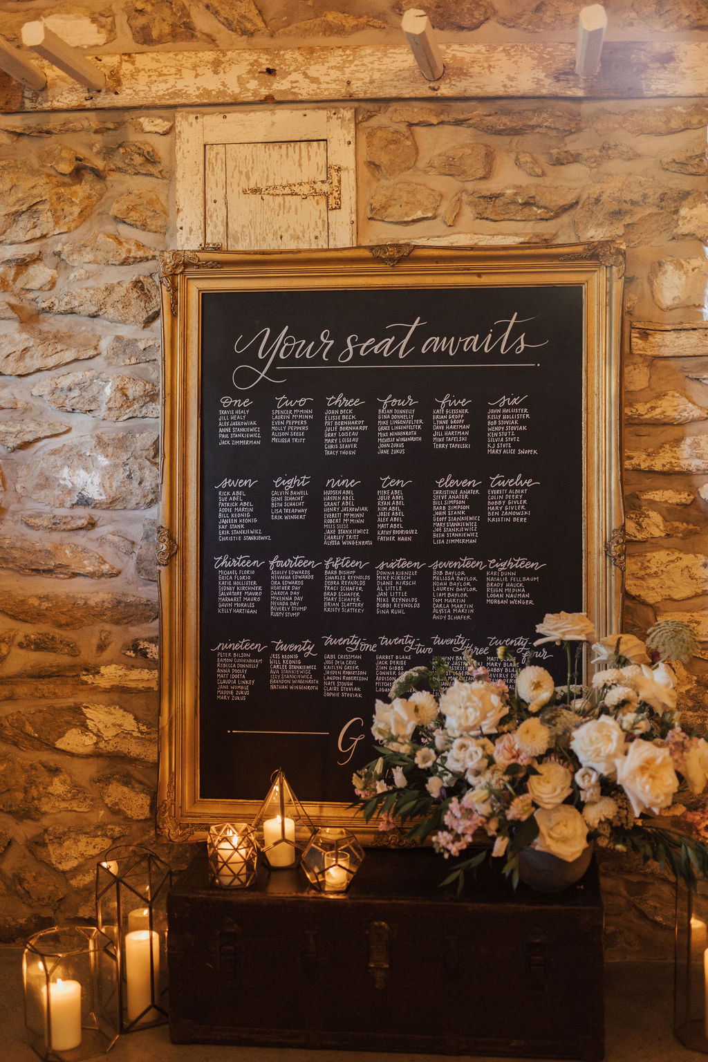 Large chalkboard with gold ornate frame used for seating chart set beside romantic candles and florals
