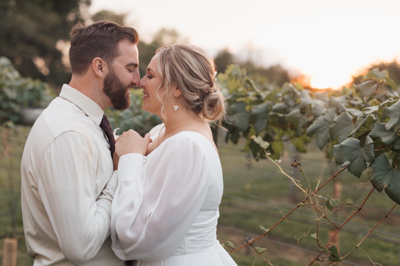 Bride and groom nose-to-nose holding hands with their eyes closed and smiling in a vineyard