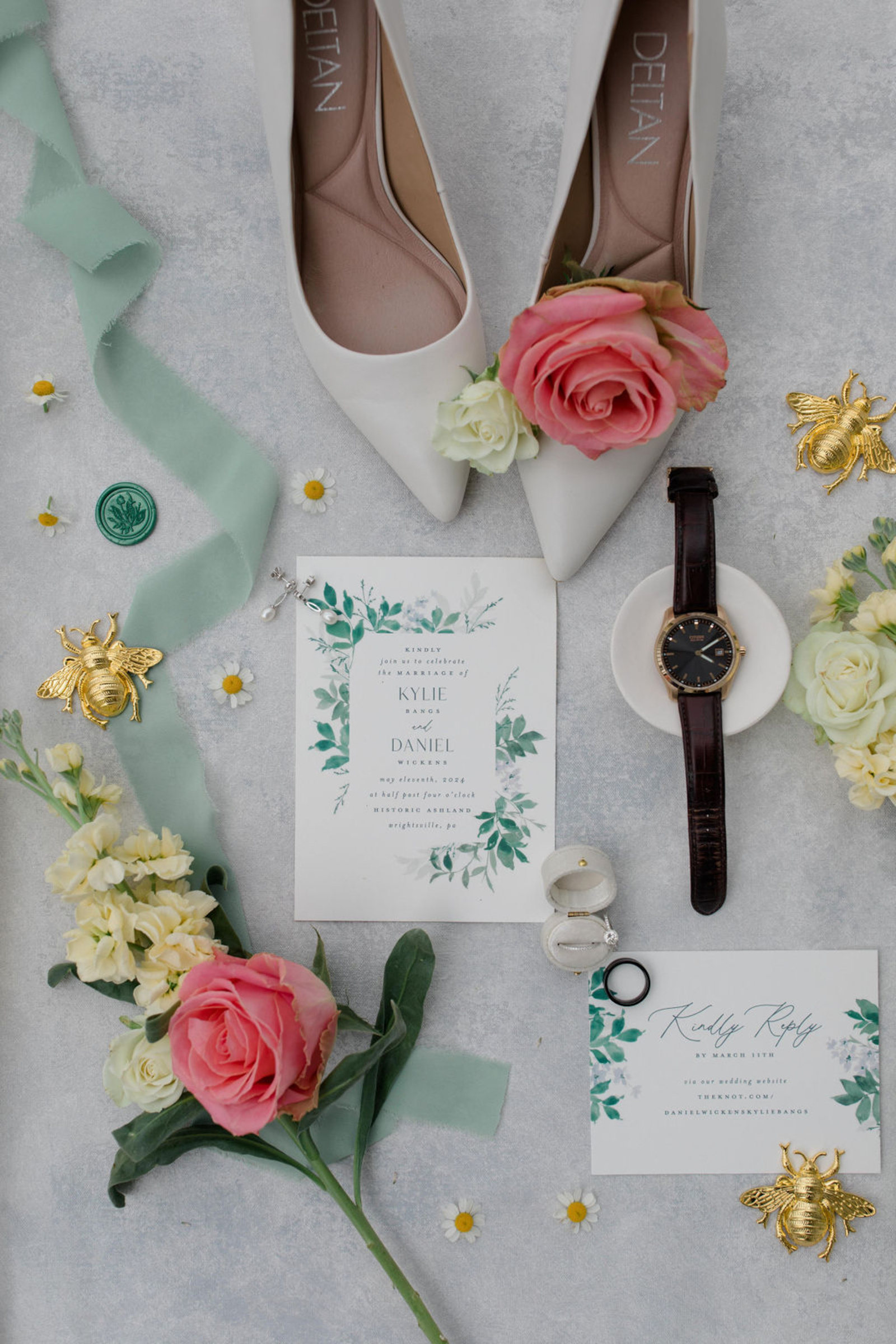 Wedding flat lay featuring bridal shoes, invitation suite, ribbon, watch, and florals styled on a light textured background.