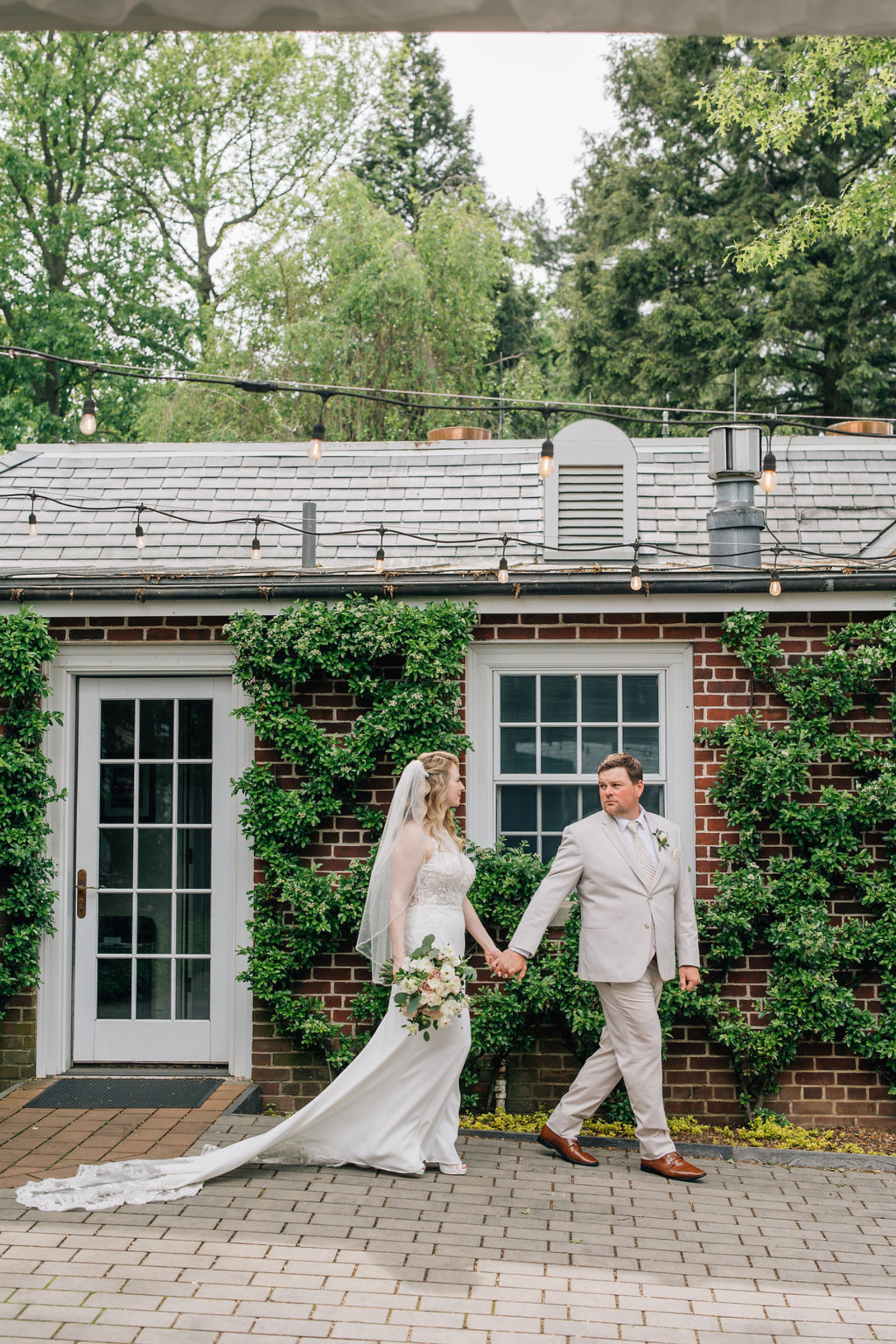 Bride and groom walking past a brick building with greenery and string lights at a Lancaster PA garden wedding venue