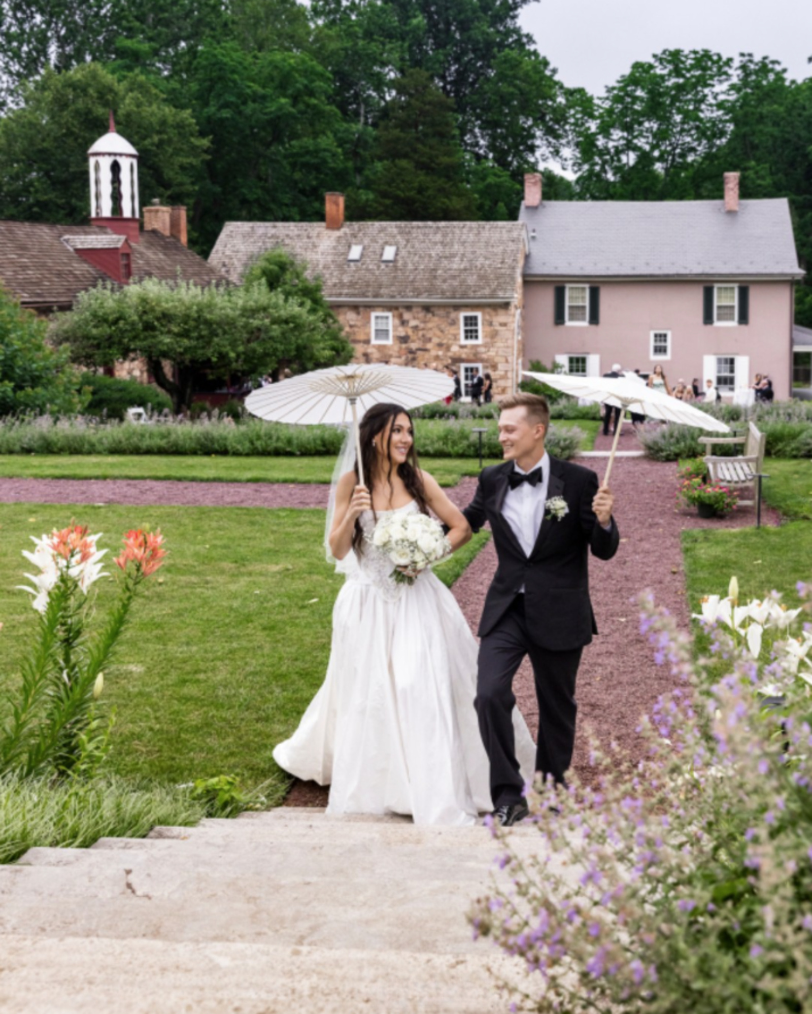 Bride and groom walking through a garden wedding venue in Lancaster PA with umbrellas, historic buildings, and landscaped grounds