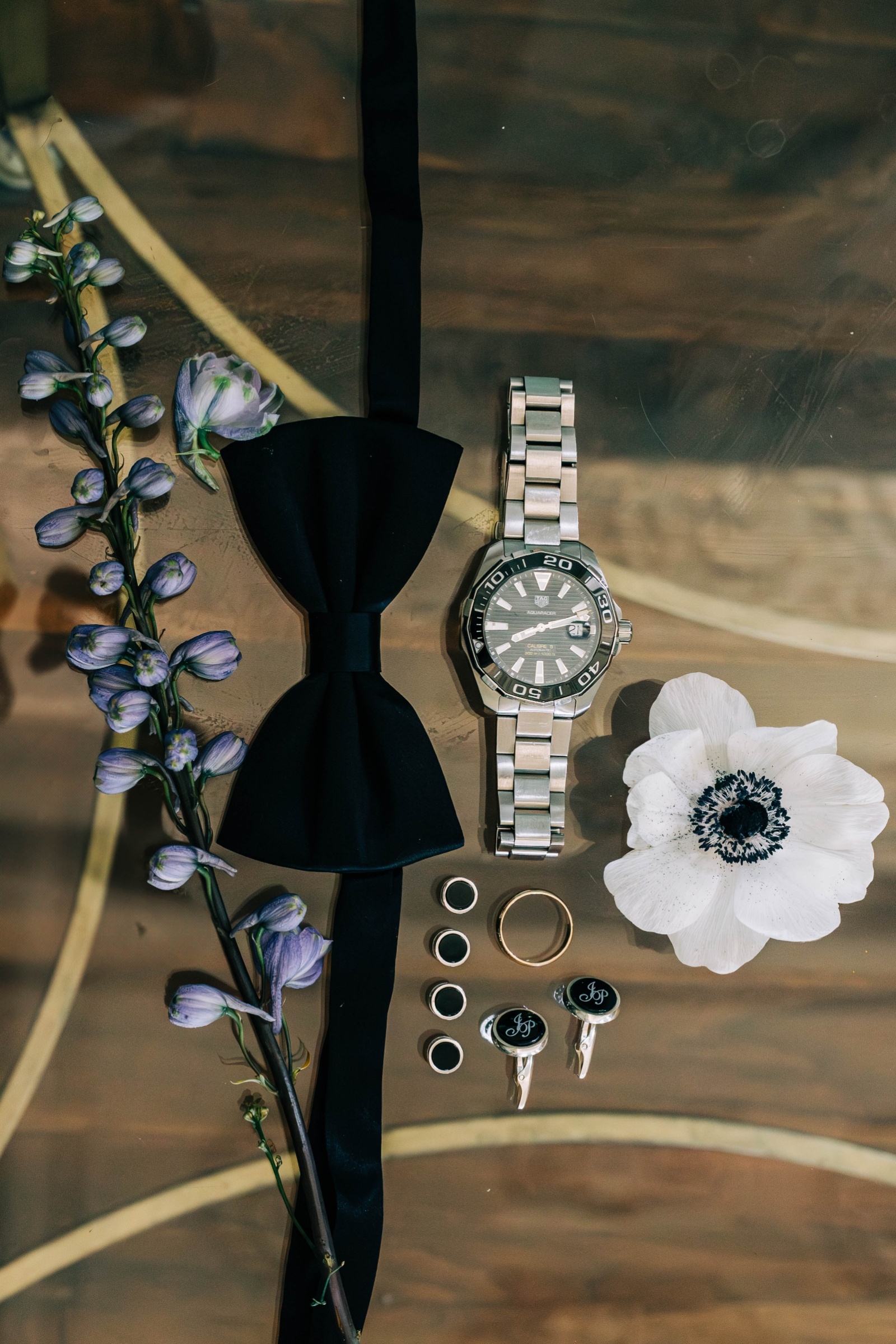 Groom wedding flat lay featuring black bow tie, watch, cufflinks, and boutonniere styled with rings and florals.