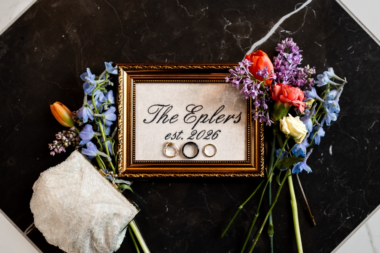 Wedding flat lay featuring gold frame with rings, vintage beaded purse, and colorful florals styled on a marble surface.