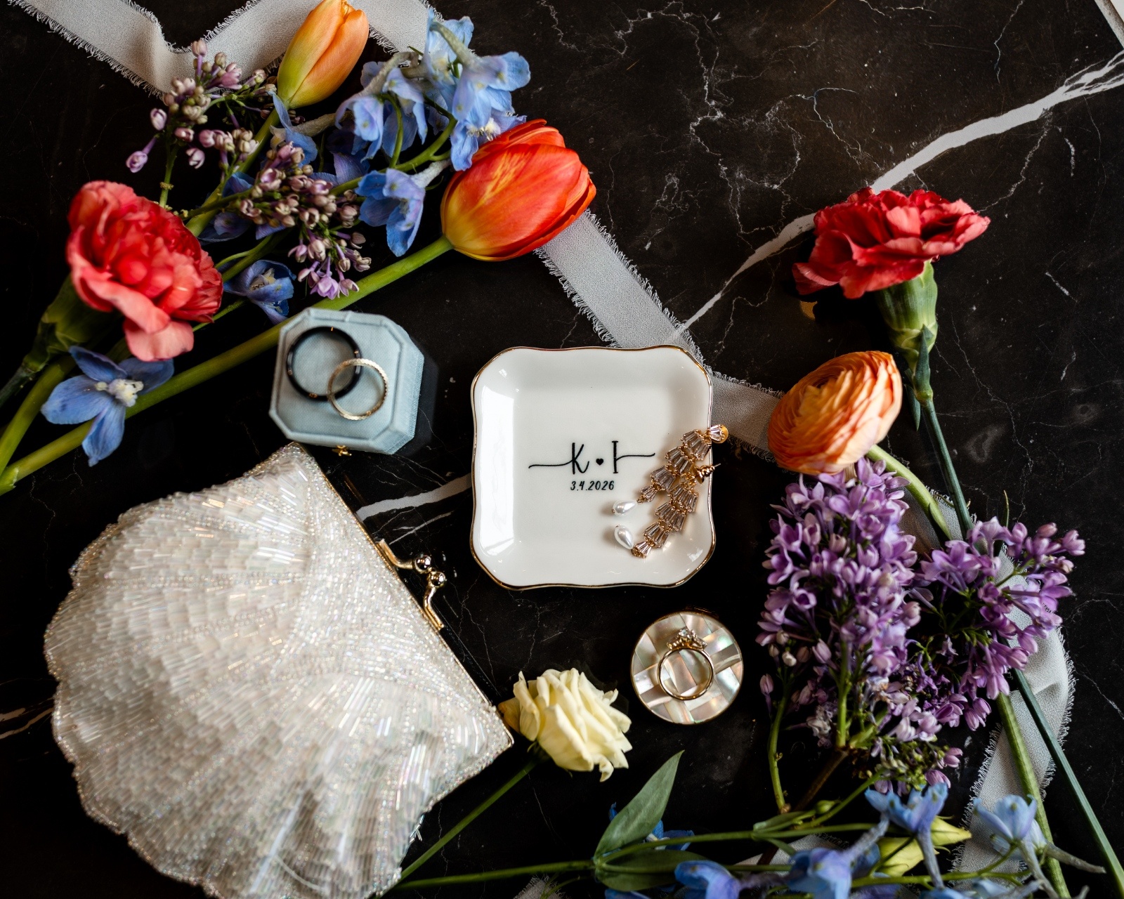Wedding flat lay with blue velvet ring box, jewelry dish, vintage purse, ribbon, and colorful florals styled on marble.