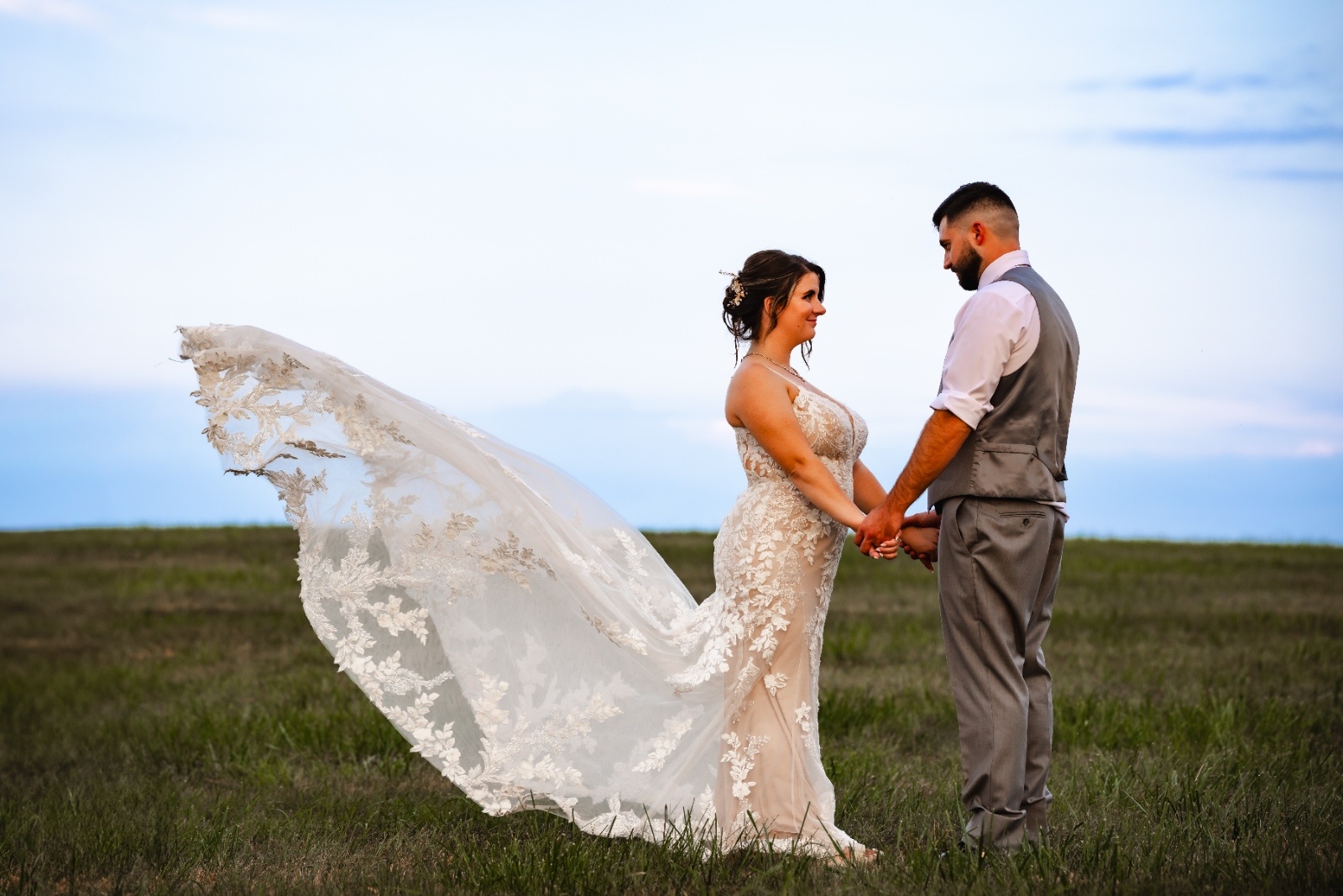 Windblown Veil Wedding Portrait Bride and groom hold hands in an open field as the bride's lace veil flows dramatically in the wind during sunset portraits.