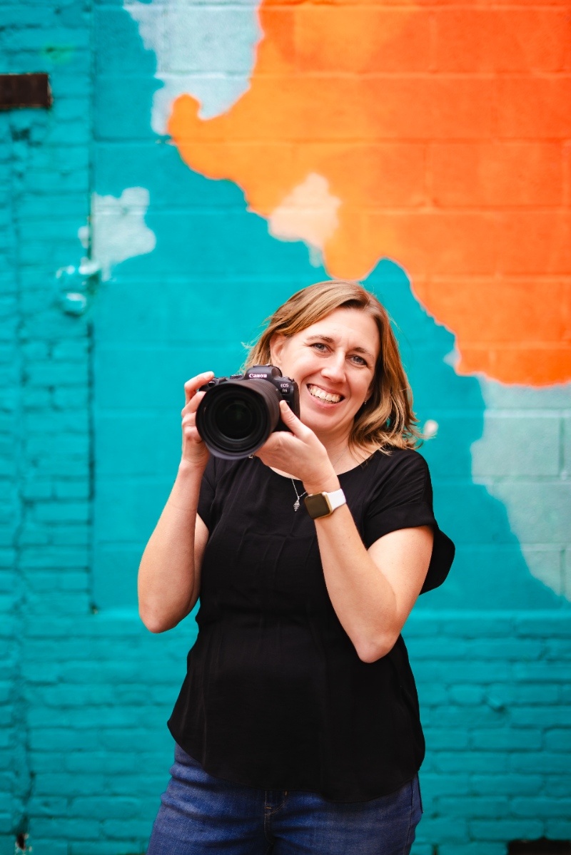 Meet Kelly Knaub of Kelly Knaub Photography Wedding photographer Kelly Knaub smiles while holding her camera in front of a colorful mural in York, Pennsylvania.