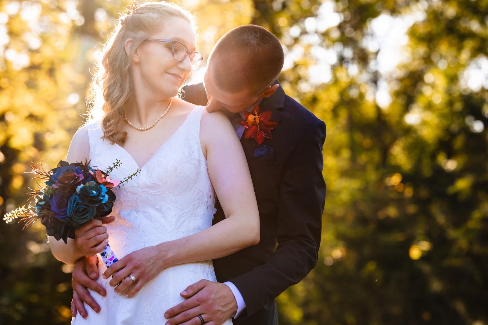 Golden Hour Wedding Portrait by Kelly Knaub Photography Groom embraces the bride from behind during a golden hour wedding portrait as she smiles and holds a colorful bouquet outdoors in Pennsylvania.