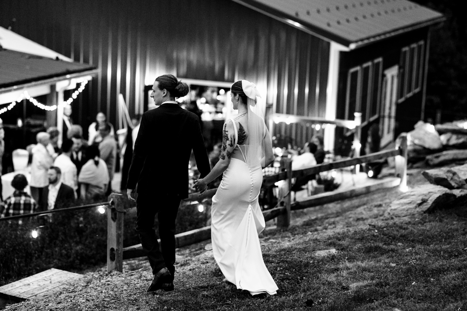Black and White Wedding Reception Portrait Newly married couple walks hand in hand toward their evening wedding reception in a timeless black and white photograph.