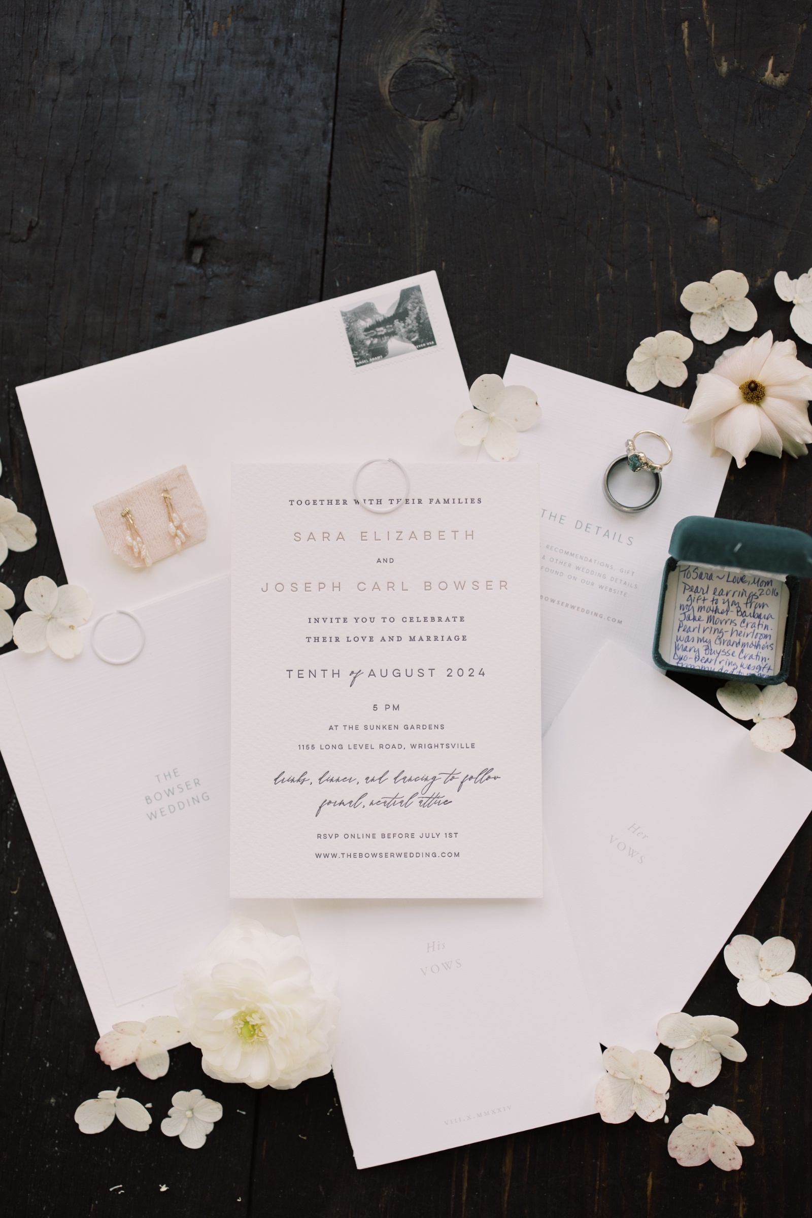 Wedding flat lay with invitation suite, vow books, engagement ring, earrings, and white florals styled on a dark wood surface.