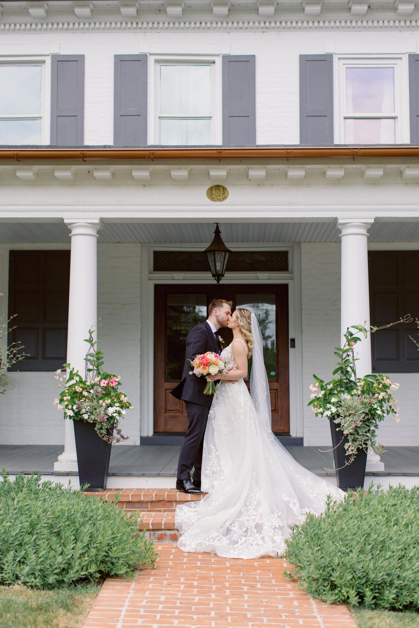 Romantic Early Summer Wedding Portrait at Historic Ashland in Pennsylvania Bride and groom kissing on the front porch of Historic Ashland in Central Pennsylvania, surrounded by soft summer florals and lush greenery during their romantic early summer wedding.