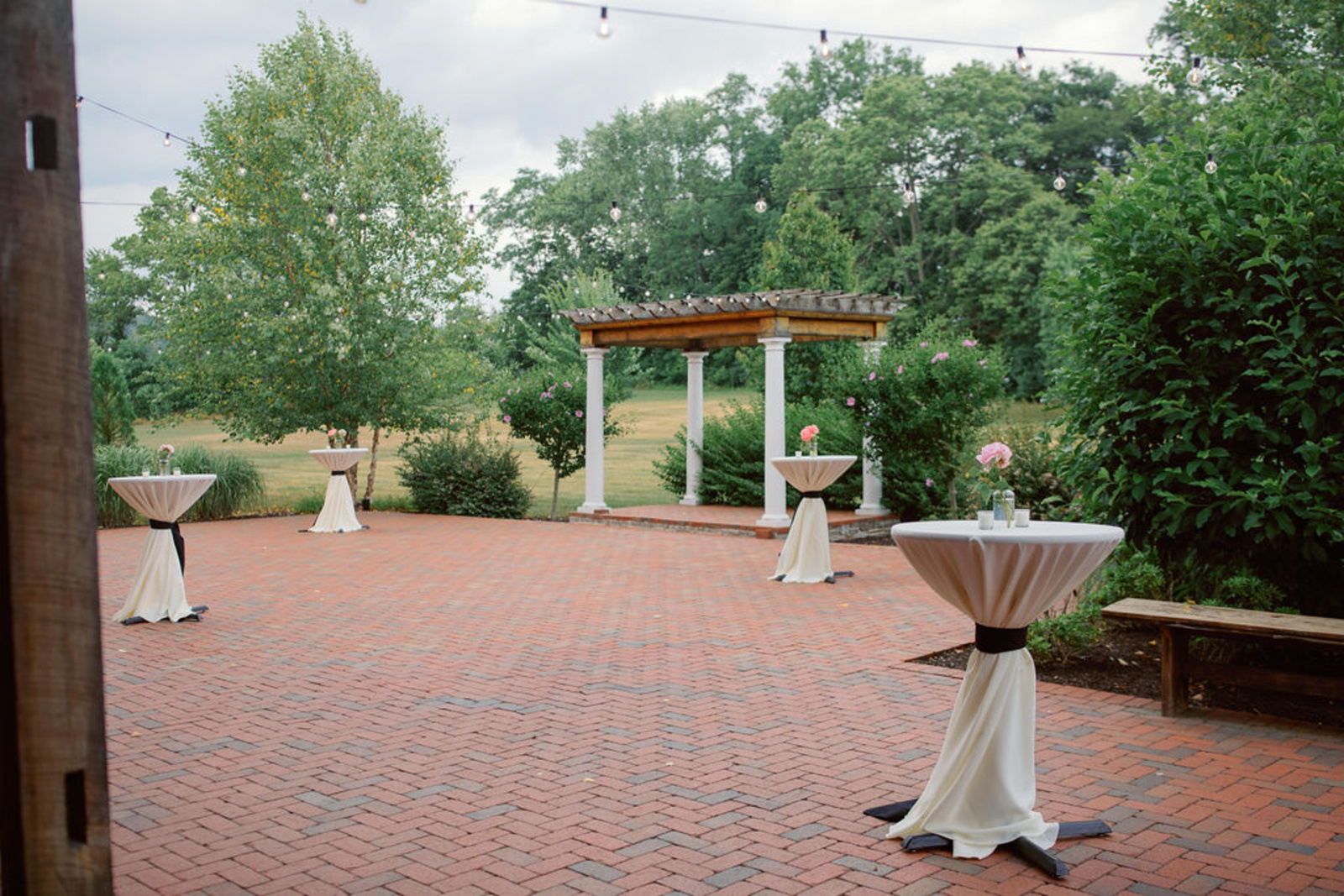Outdoor Cocktail Hour Space at Historic Ashland Brick courtyard cocktail hour space at Historic Ashland featuring string lights, greenery, and cocktail tables before a summer wedding reception.