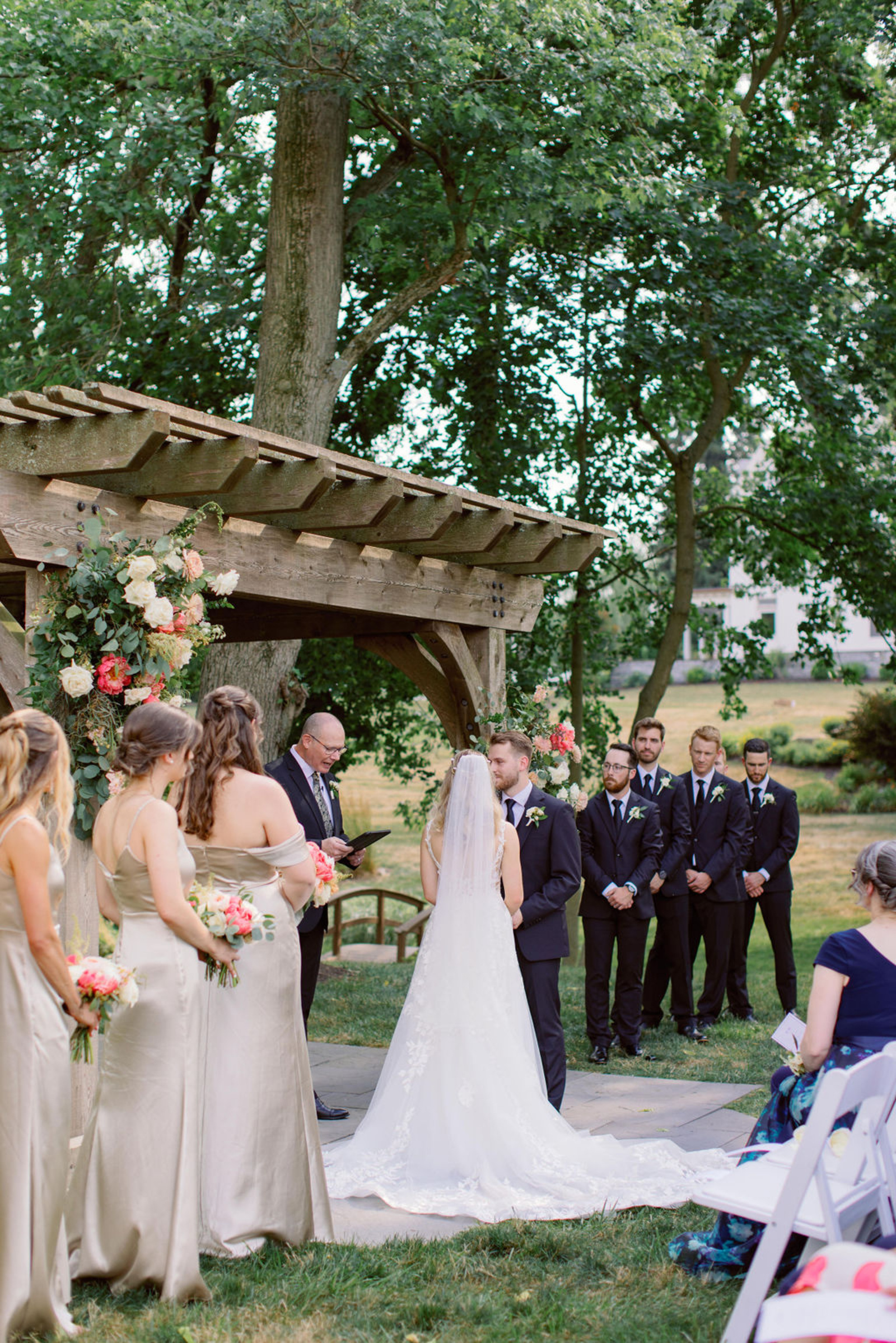 Outdoor Wedding Ceremony at Historic Ashland Bride and groom exchanging vows beneath a wooden pergola decorated with summer florals during an outdoor wedding ceremony at Historic Ashland.