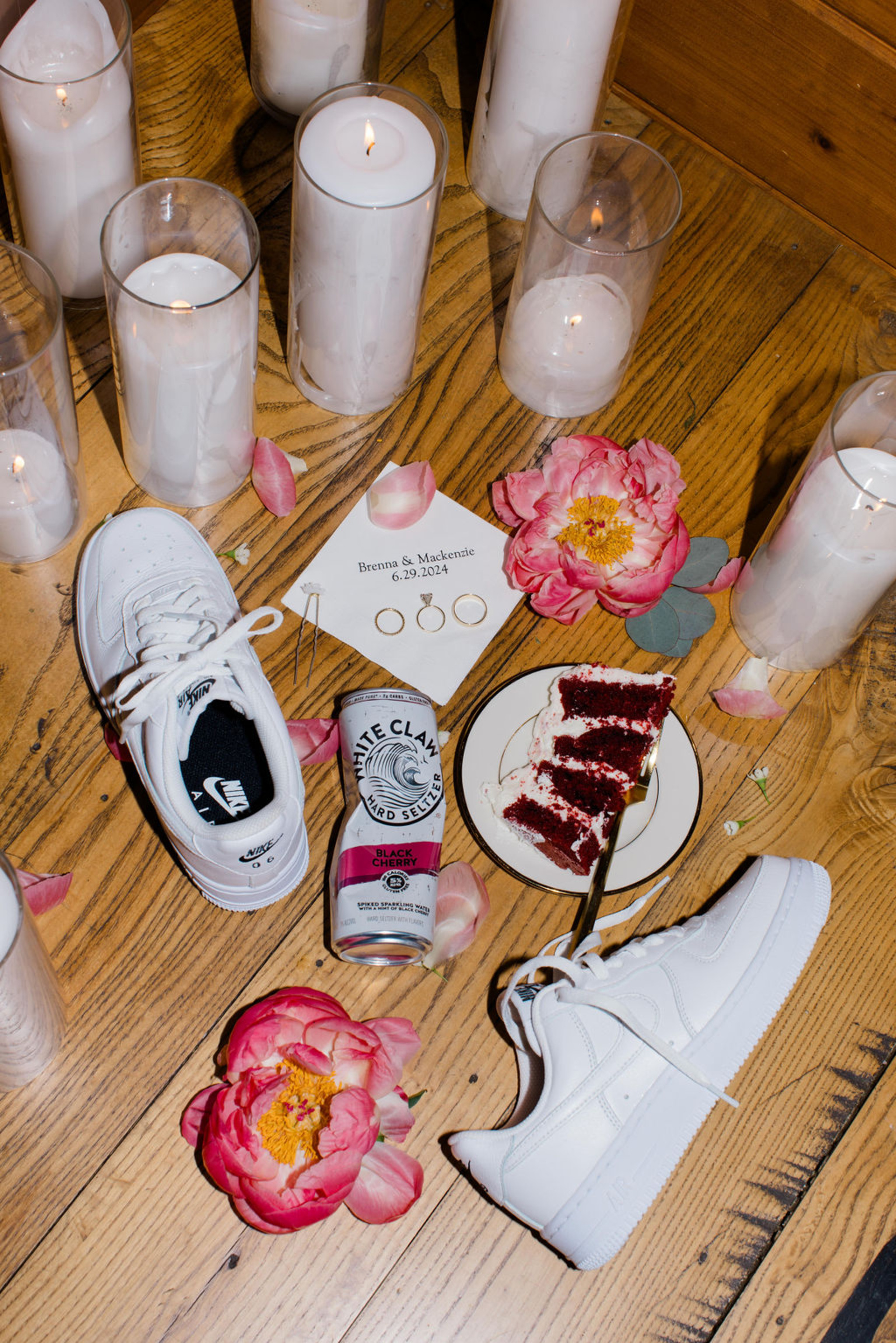 Fun End of the Night Wedding Detail Flat Lay at Historic Ashland End of the night wedding flat lay featuring white Nike sneakers, pink florals, wedding rings, candles, cake, and a White Claw on a wood floor.