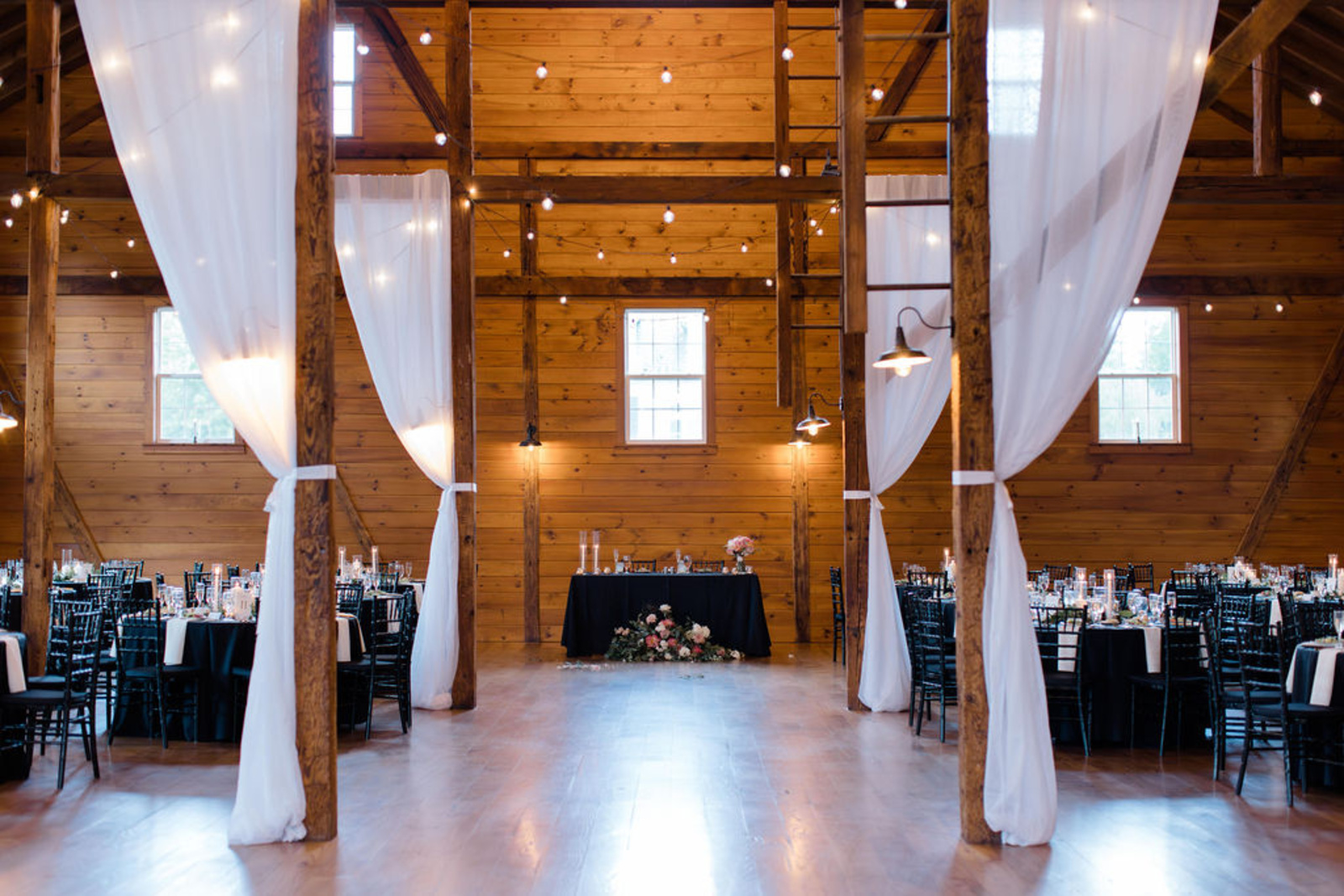 Historic Ashland Lodge Reception Space Romantic barn reception space at Historic Ashland with wood beams, draped white fabric, string lights, and black-and-white reception tables.