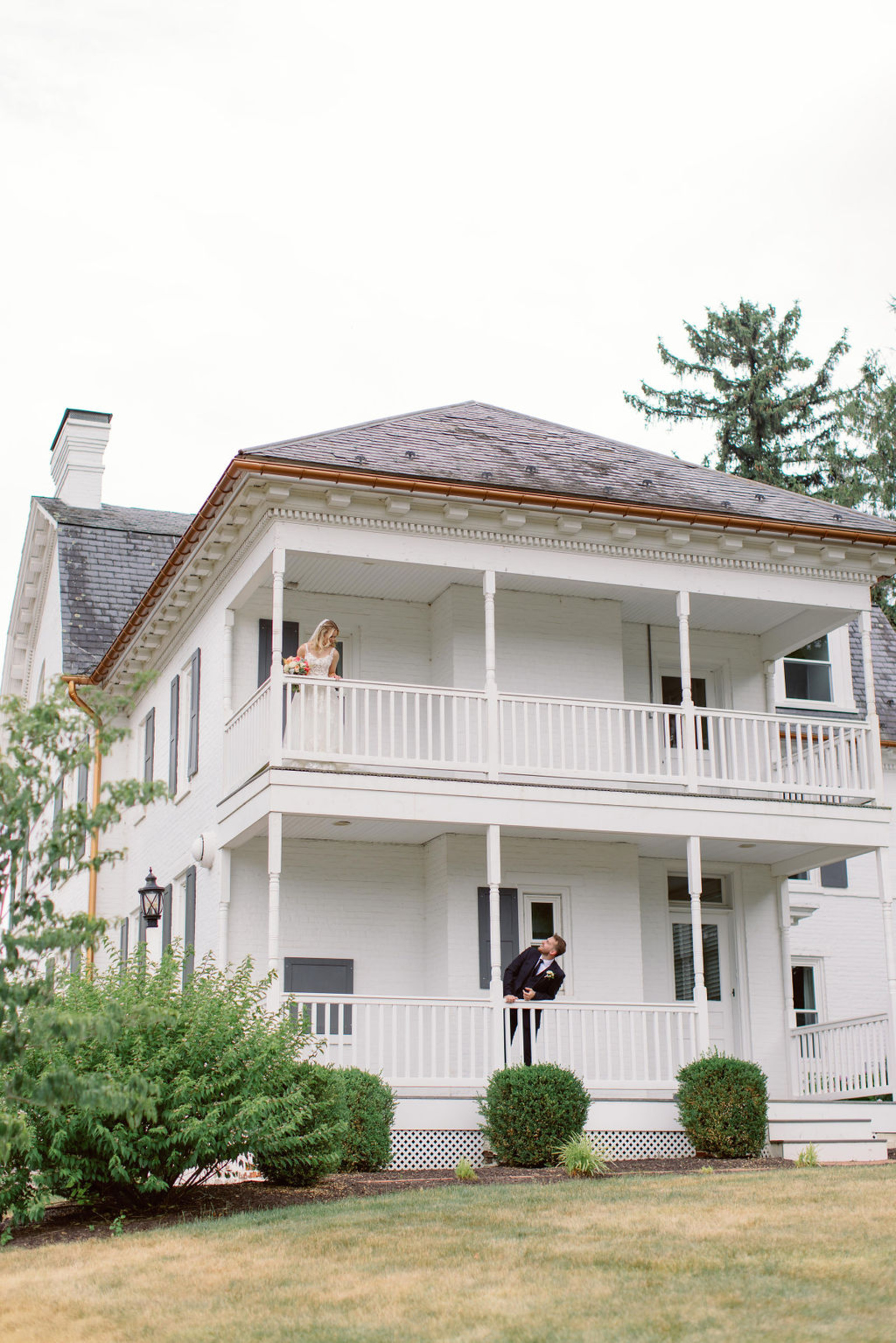 Bride and Groom Portrait at Historic Ashland Bride standing on the second-story balcony while the groom looks up from the porch below at Historic Ashland during their early summer Pennsylvania wedding.