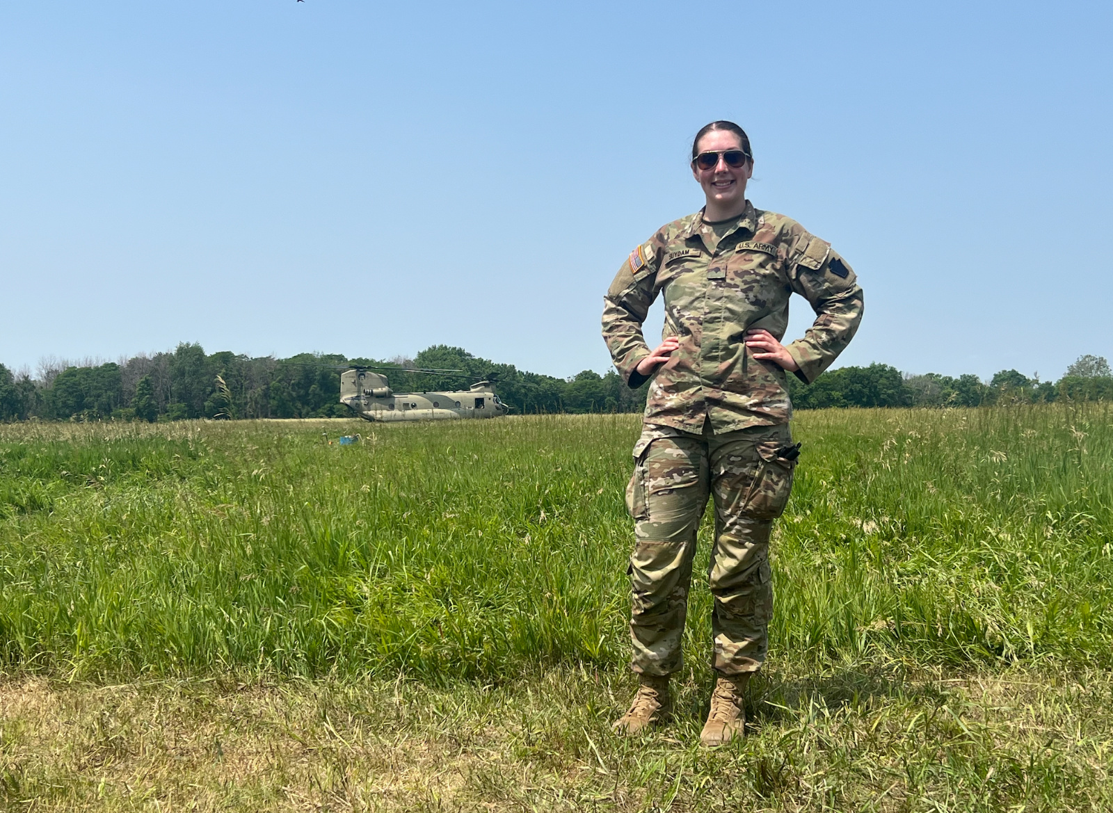 Kelly Suydam, owner of Sips & Shakers Mobile Bartending, standing in uniform on a grassy field with a military helicopter in the background, representing her service as a Sergeant in the Pennsylvania Army National Guard.