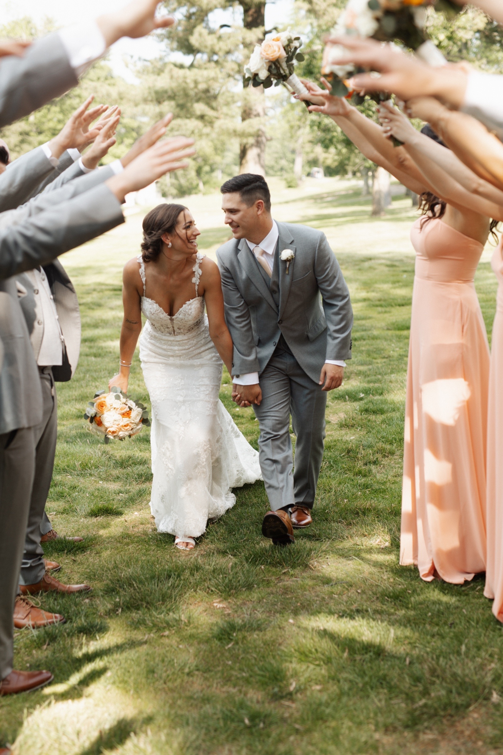 Bride and groom smiling as they walk hand in hand through a tunnel formed by their wedding party, with bridesmaids in peach dresses and groomsmen in gray suits raising bouquets in celebration.