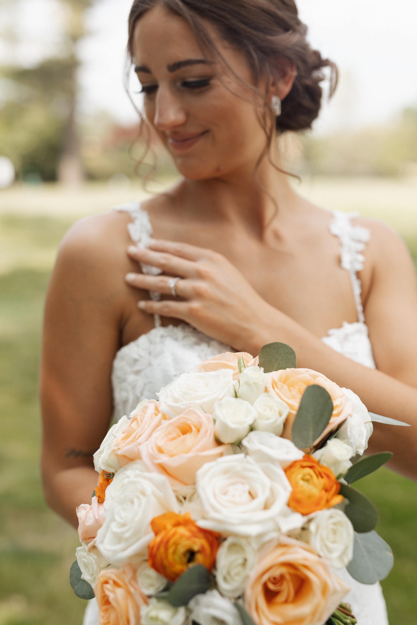 Bride holding a bouquet of peach, white, and orange roses while gently touching her shoulder strap, smiling softly with her hair in a loose updo and wearing a lace wedding gown.