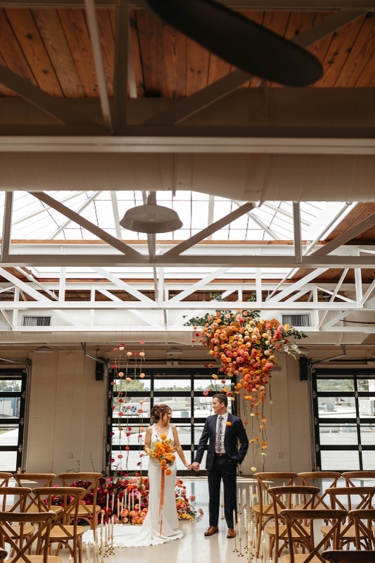 Bold Hanging Floral Installation for Modern Wedding Decor Dramatic hanging floral installation with bold orange and warm-toned blooms inside a modern wedding venue, highlighting statement floral trends for 2026 weddings.