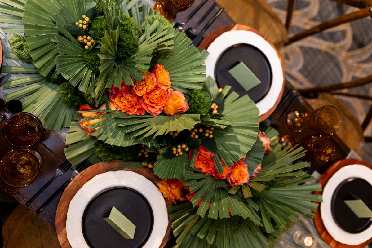 A top-down view of a wedding table featuring lush greenery, palm fans, and bright orange roses arranged in a bold Oklahoma!-inspired centerpiece.