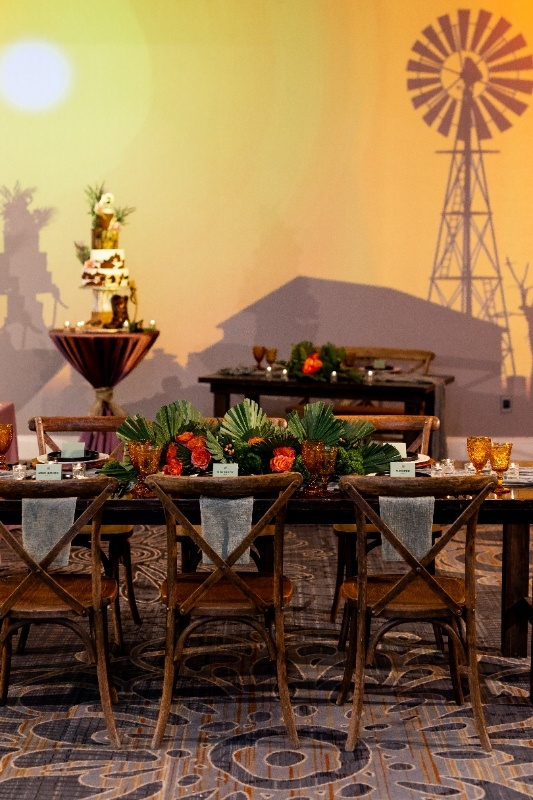 A rustic wedding reception table with amber glasses, greenery, and bold orange florals is set beneath warm lighting and a backdrop featuring a windmill silhouette.