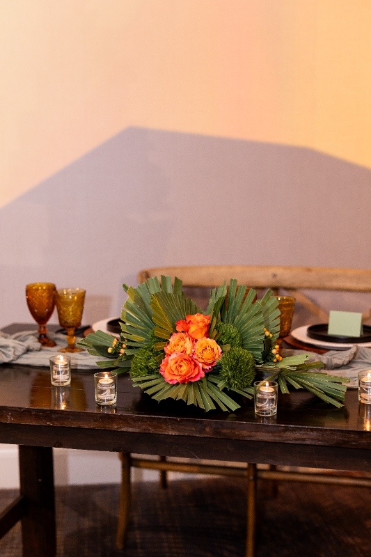 A wedding centerpiece made of green palms, orange roses, and soft candlelight adds western elegance to a rustic sweetheart table