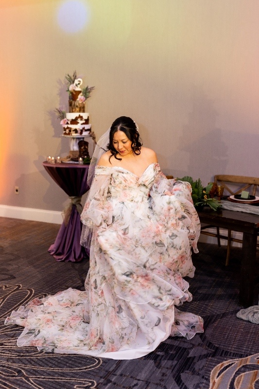 A bride twirls in a flowing floral off-the-shoulder gown during a western-inspired styled wedding shoot, with a rustic cake table and warm lighting in the background.
