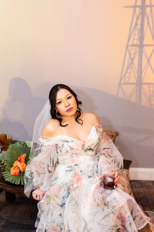 A bride in a floral gown and veil sits gracefully, holding a drink and embodying effortless western charm against a rustic Oklahoma!-inspired backdrop.