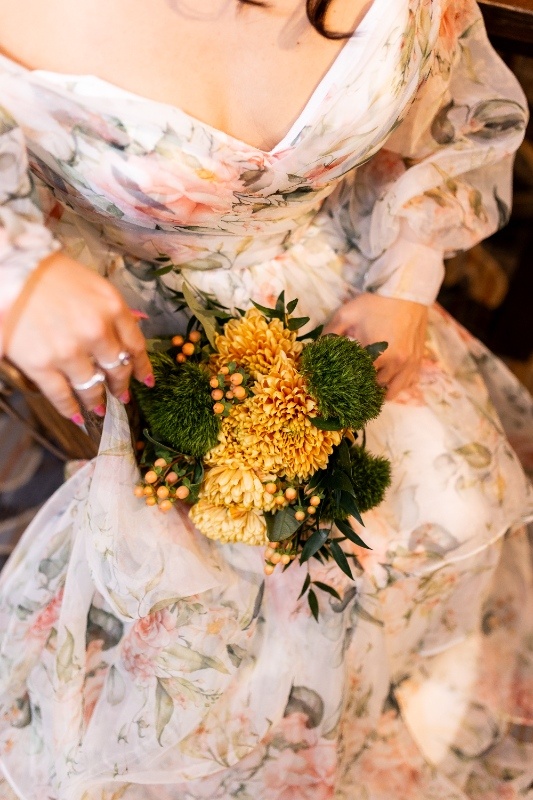 Close-up of a bride in a floral off-the-shoulder gown holding a bouquet of yellow mums, green moss balls, and berry accents, capturing the warm rustic tones of this Oklahoma!-inspired wedding shoot.