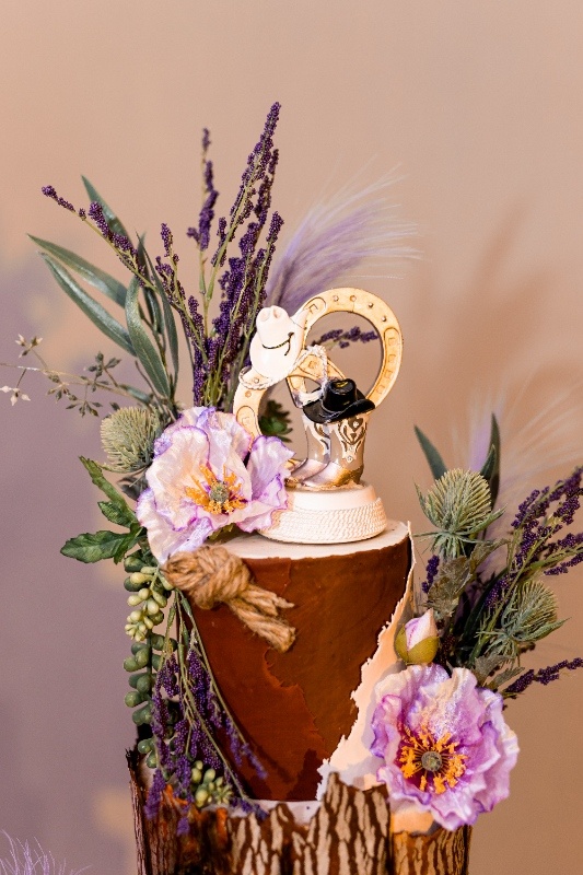 Close-up of a rustic cake topper featuring a gold horseshoe, cowboy boots, and hats surrounded by lavender sprigs and soft purple florals, perfect for a western-themed wedding.