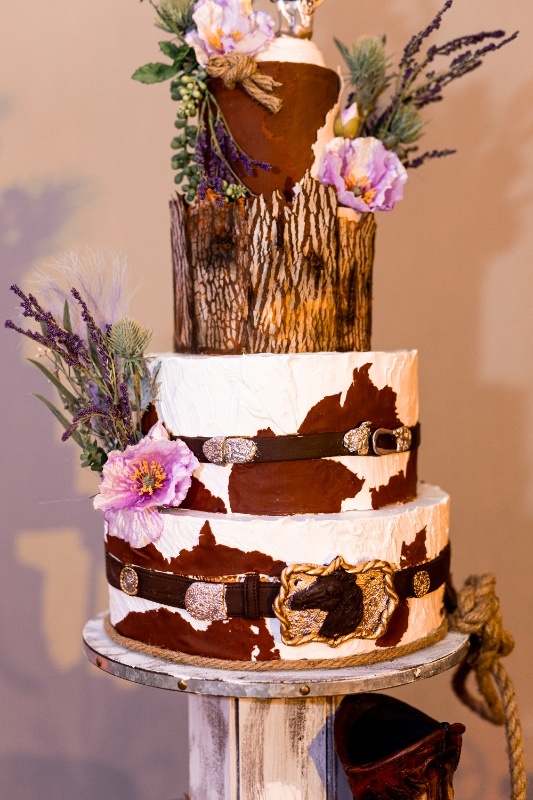 A three-tiered wedding cake featuring cowhide patterns, leather belt accents, and floral embellishments, blending rustic western charm with creative Oklahoma!-inspired design.