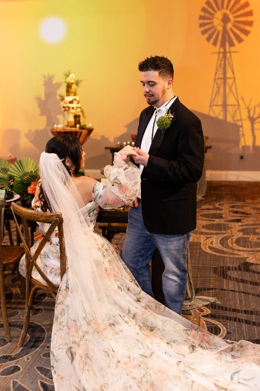 The bride in a flowing floral gown and the groom in jeans and a blazer share a sweet moment under warm lighting, surrounded by rustic decor and a windmill backdrop inspired by Oklahoma!
