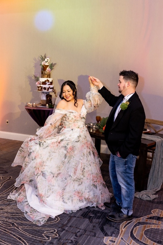 The bride twirls in a flowing floral gown while the groom in a casual western look smiles beside her, surrounded by rustic décor and warm lighting inspired by Oklahoma!.