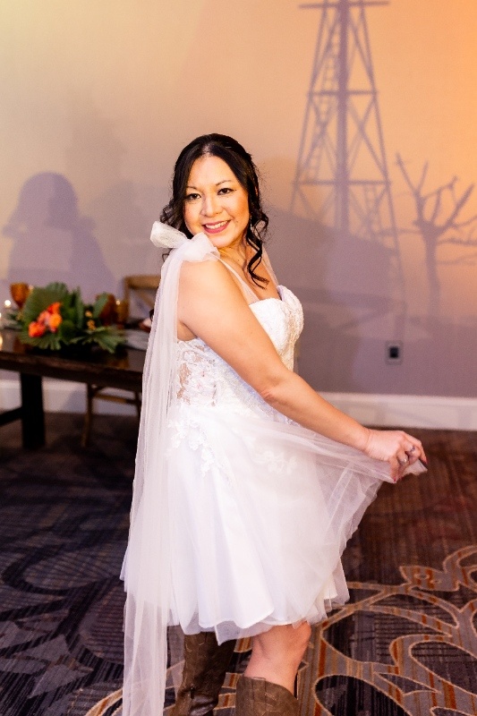 A bride smiles in a short lace wedding dress paired with a long veil and cowboy boots, embodying playful country charm in this Oklahoma!-inspired styled shoot.