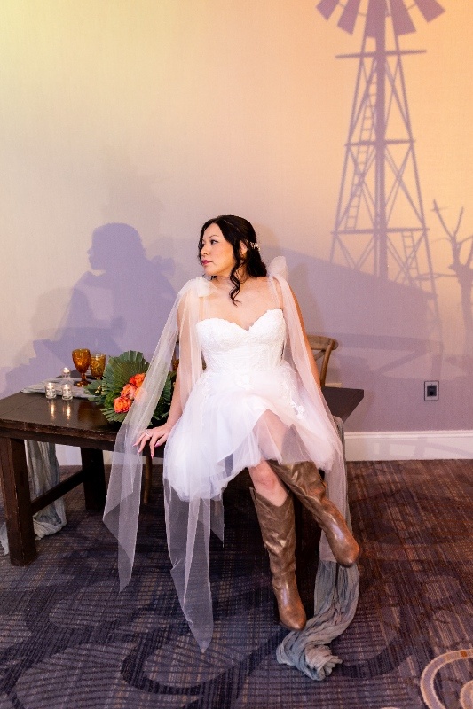 The bride sits gracefully in a short lace gown and tall brown boots against a rustic backdrop, highlighting the fun and flirty side of Oklahoma! wedding inspiration.