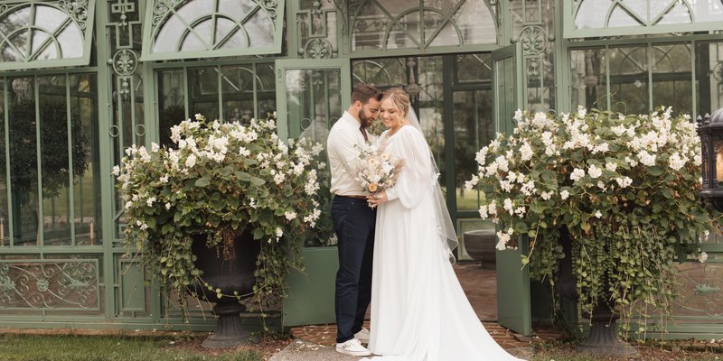 Bride and groom standing in front of a greenhouse surrounded by white flowers during a romantic garden wedding