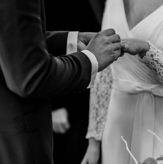 Black and white photo of a couple exchanging rings during an intimate self-uniting wedding ceremony, featuring a bride in a long sleeve lace wedding dress and a close-up of the couple’s hands.