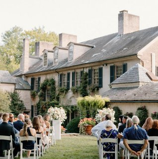 Outdoor garden wedding ceremony at a historic estate in Lancaster, PA with guests seated on white chairs in front of a brick manor surrounded by greenery and florals