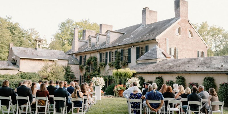 Outdoor garden wedding ceremony at a historic estate in Lancaster, PA with guests seated on white chairs in front of a brick manor surrounded by greenery and florals