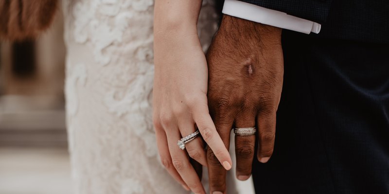 Bride and groom holding hands during a wedding ceremony, featuring close-up wedding rings and lace bridal gown details.