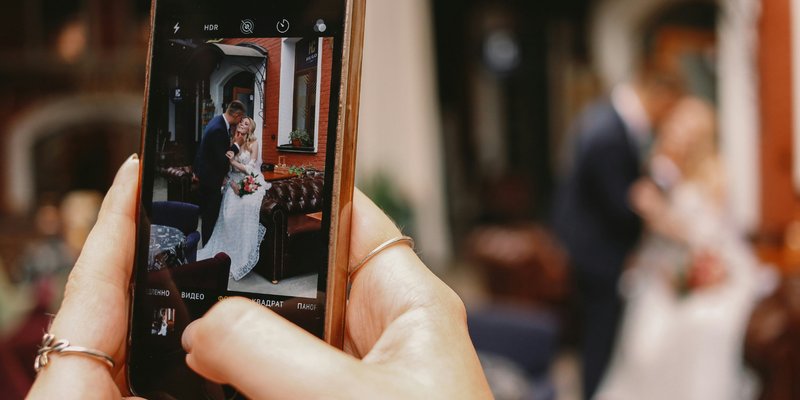 Guest photographing a newlywed couple on a smartphone, illustrating how couples can collect candid wedding photos and videos through QR code photo sharing.