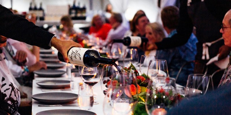 Server pours red wine at a long rehearsal dinner table decorated with colorful floral centerpieces as wedding guests gather to celebrate before the wedding day.